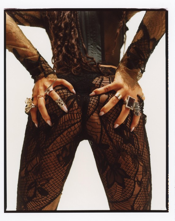 A woman in a black lace outfit with rings on her fingers poses with her hands on her hips.