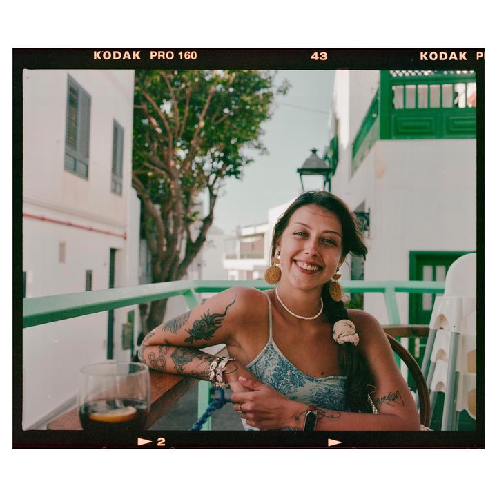 A smiling woman with tattoos on her arms is sitting at a table with a drink, with white buildings and green railings in the background.
