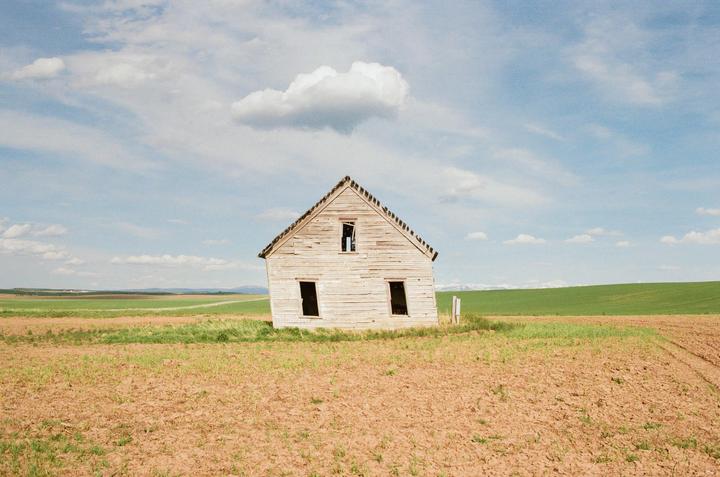 An abandoned house stands in a field under a partly cloudy sky.