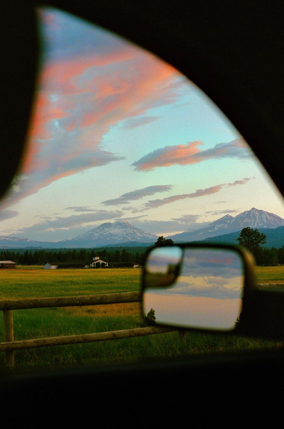A scenic view of a rural landscape with mountains and a sunset sky is seen through a car window and reflected in a side mirror.