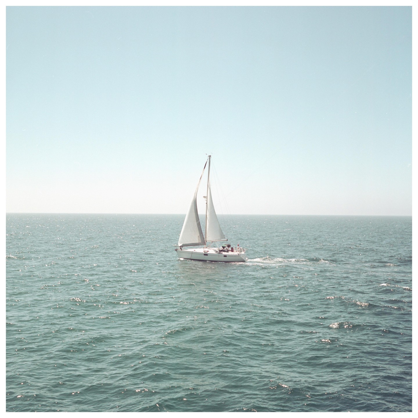 A sailboat with white sails moves across a vast expanse of water under a clear sky.
