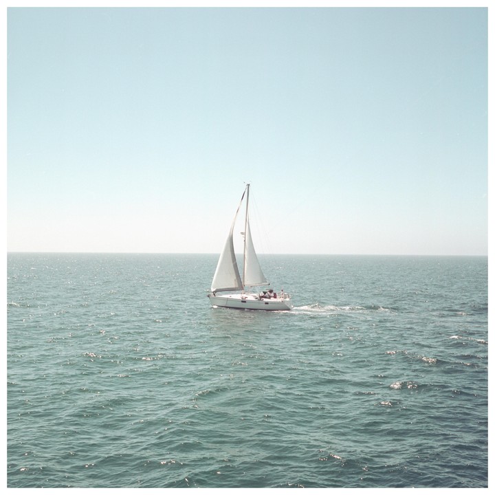A sailboat with white sails moves across a vast expanse of water under a clear sky.