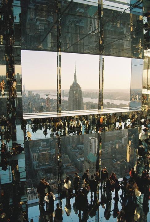 A crowd of people stands on a mirrored floor and looks out at the New York City skyline with the Empire State Building prominent in the center.