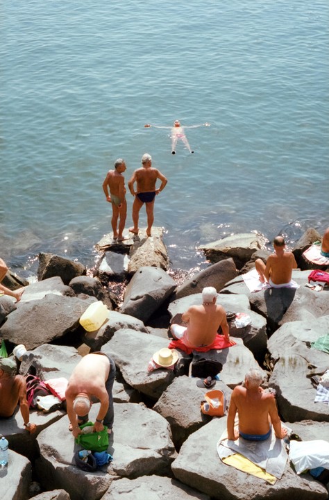 Several men are relaxing on large rocks by the water, with one man floating on his back in the sea.