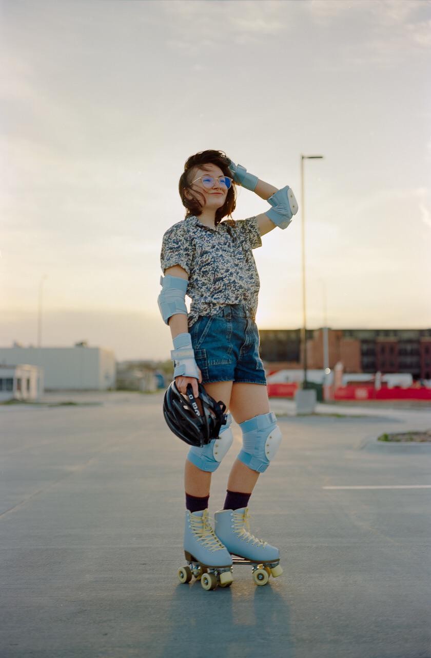 A woman wearing roller skates and protective gear stands in a parking lot, holding a helmet.