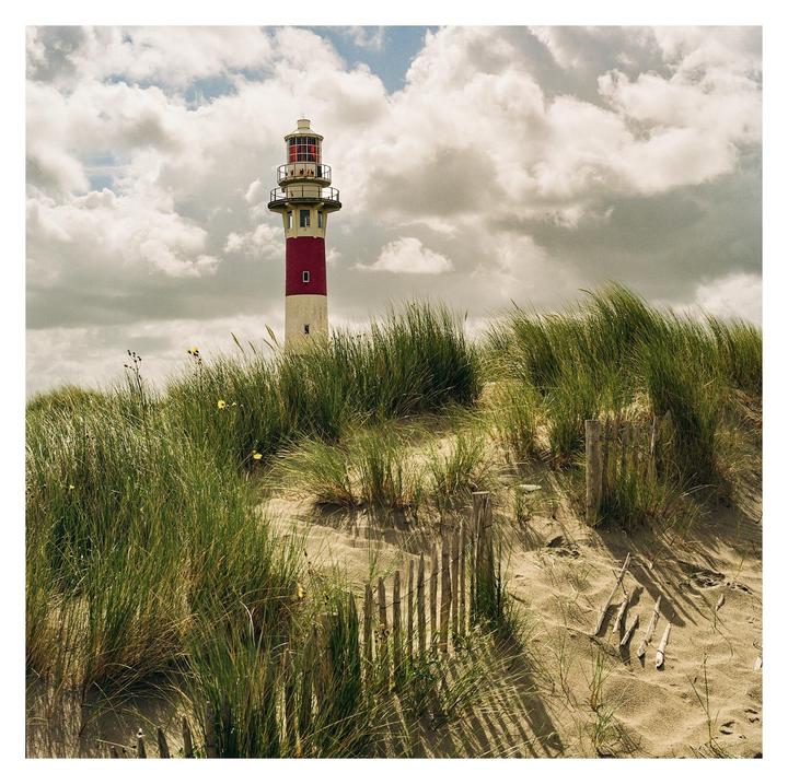 A red and white lighthouse stands behind a sandy dune covered in tall grass and a wooden fence.