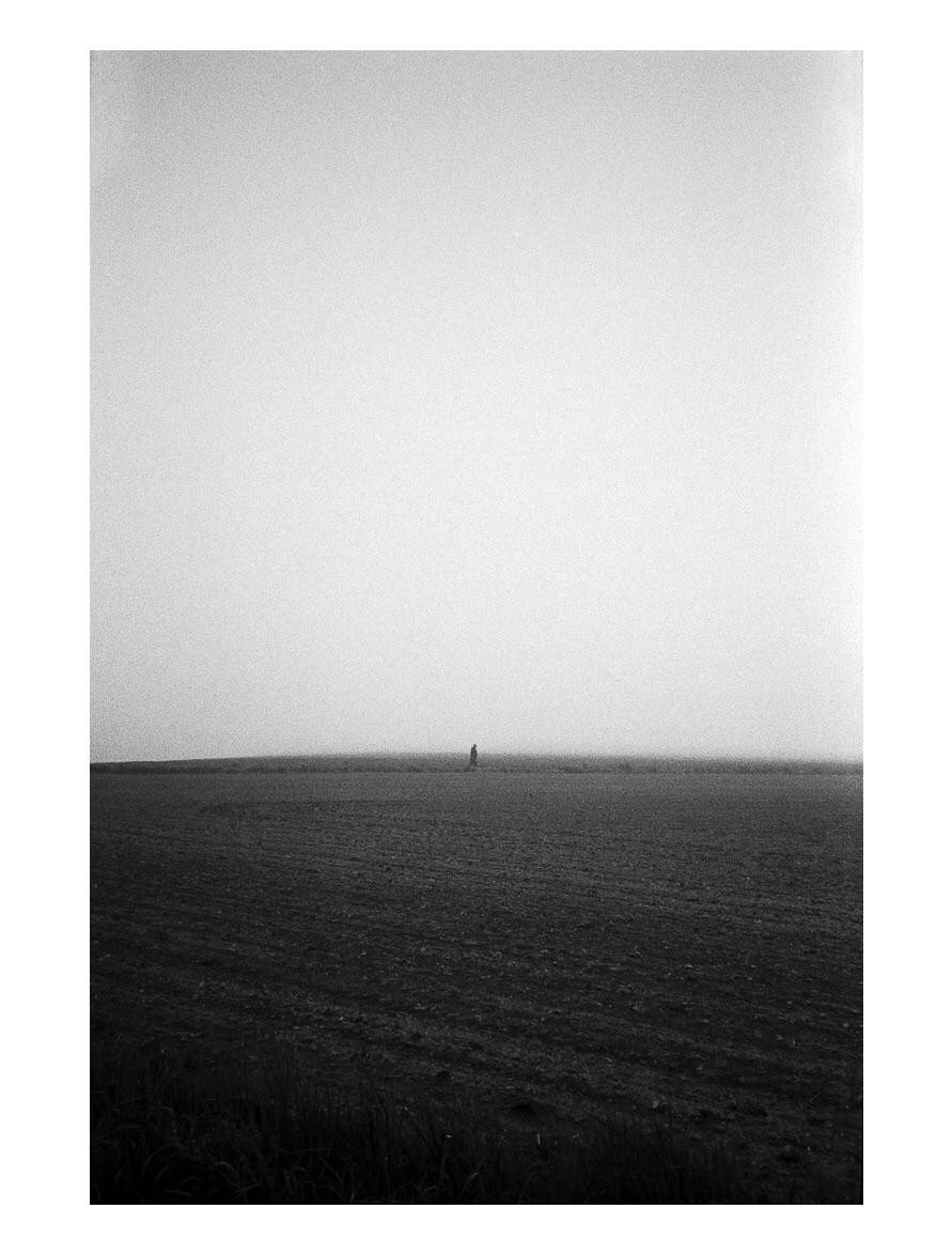 A lone man walks across a vast, tilled field under a hazy sky.