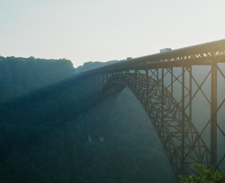 A long bridge spans a deep gorge filled with trees, with a ray of light shining through the haze.