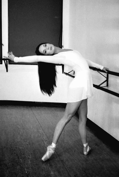 A woman in a white dress and ballet shoes leans back against a ballet barre in a studio.