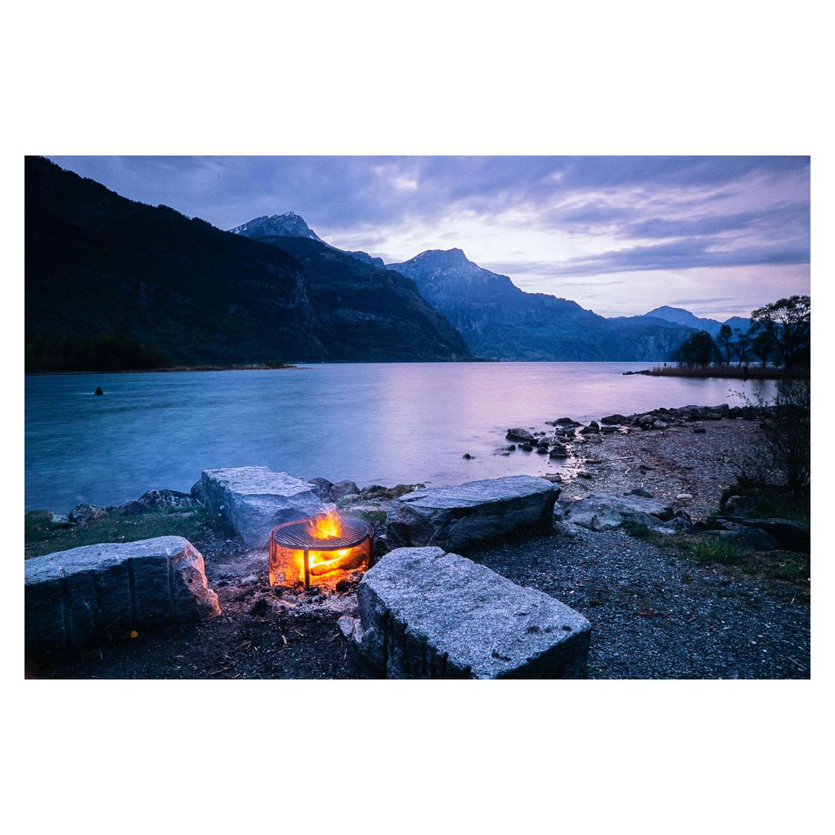 A fire pit surrounded by large rocks burns brightly on the shore of a lake with mountains in the background under a cloudy sky.