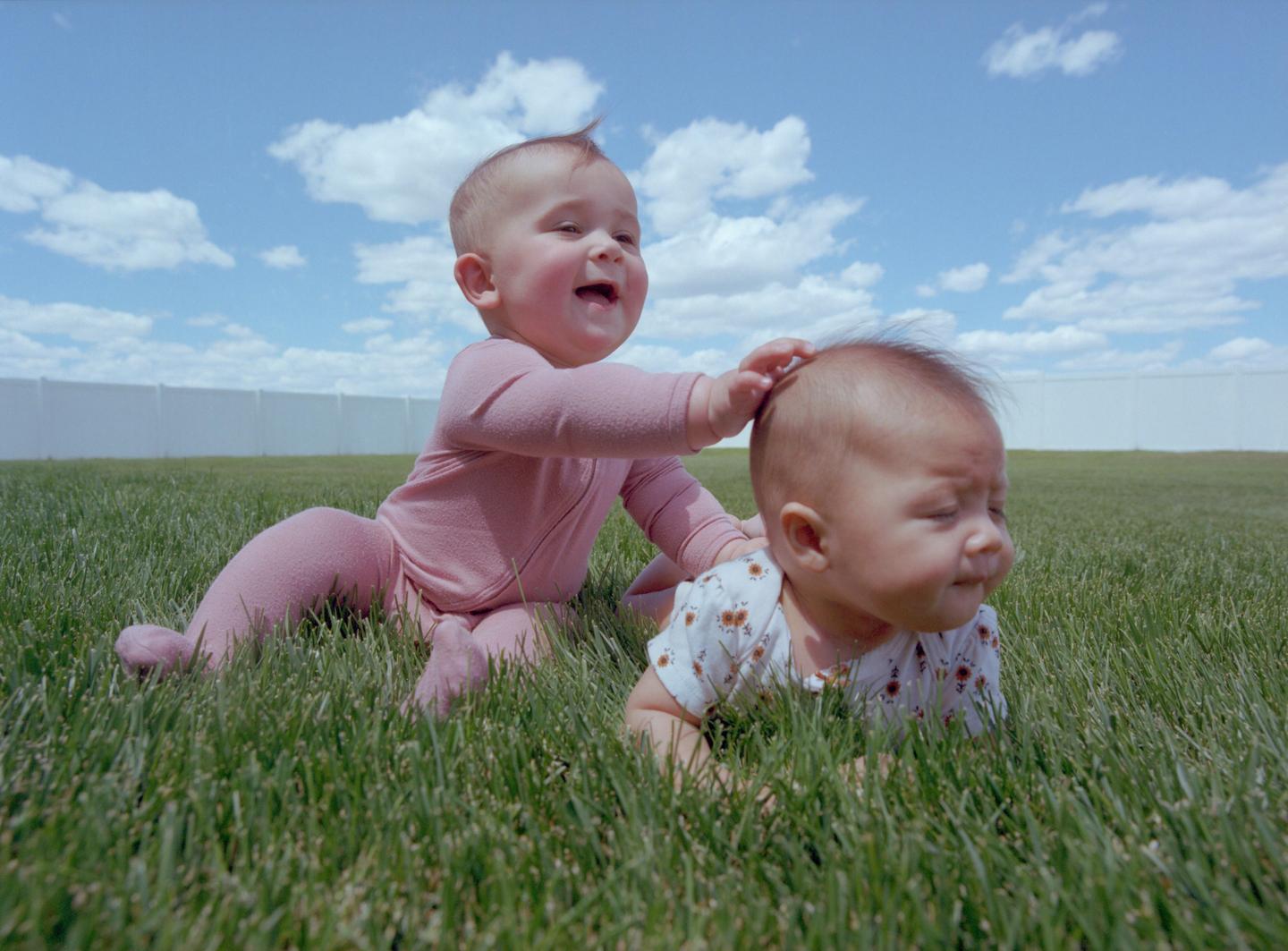 Two babies are playing in the grass under a blue sky with white clouds.