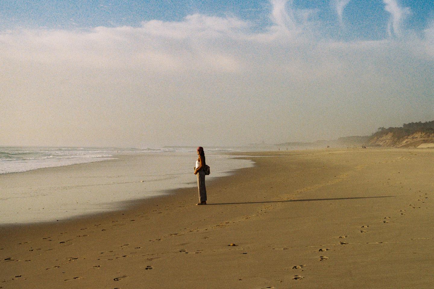 A woman stands on a sandy beach looking out at the ocean waves under a hazy sky.