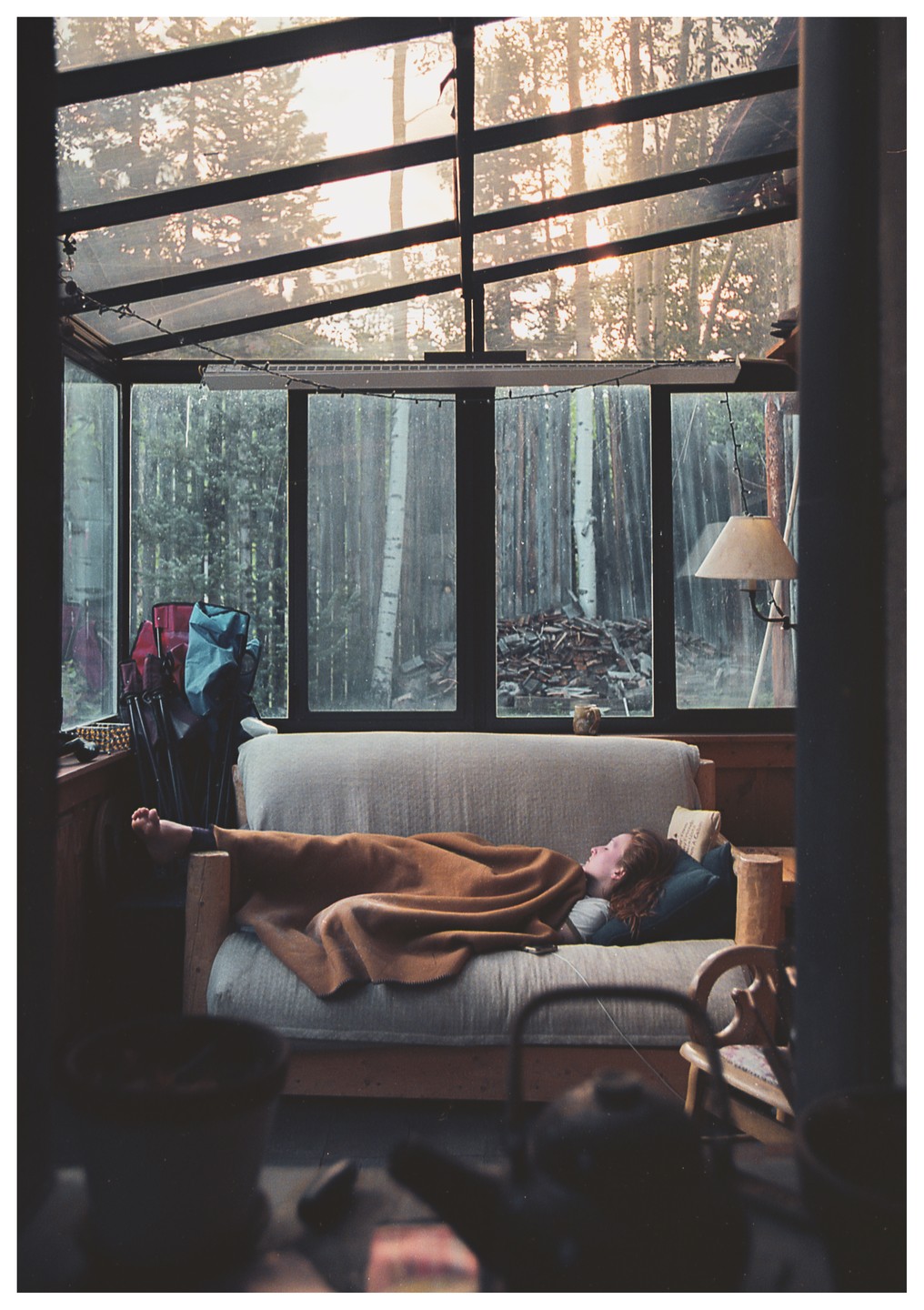 A woman sleeps on a sofa covered with a blanket in a sunroom with large windows looking out onto a forest.