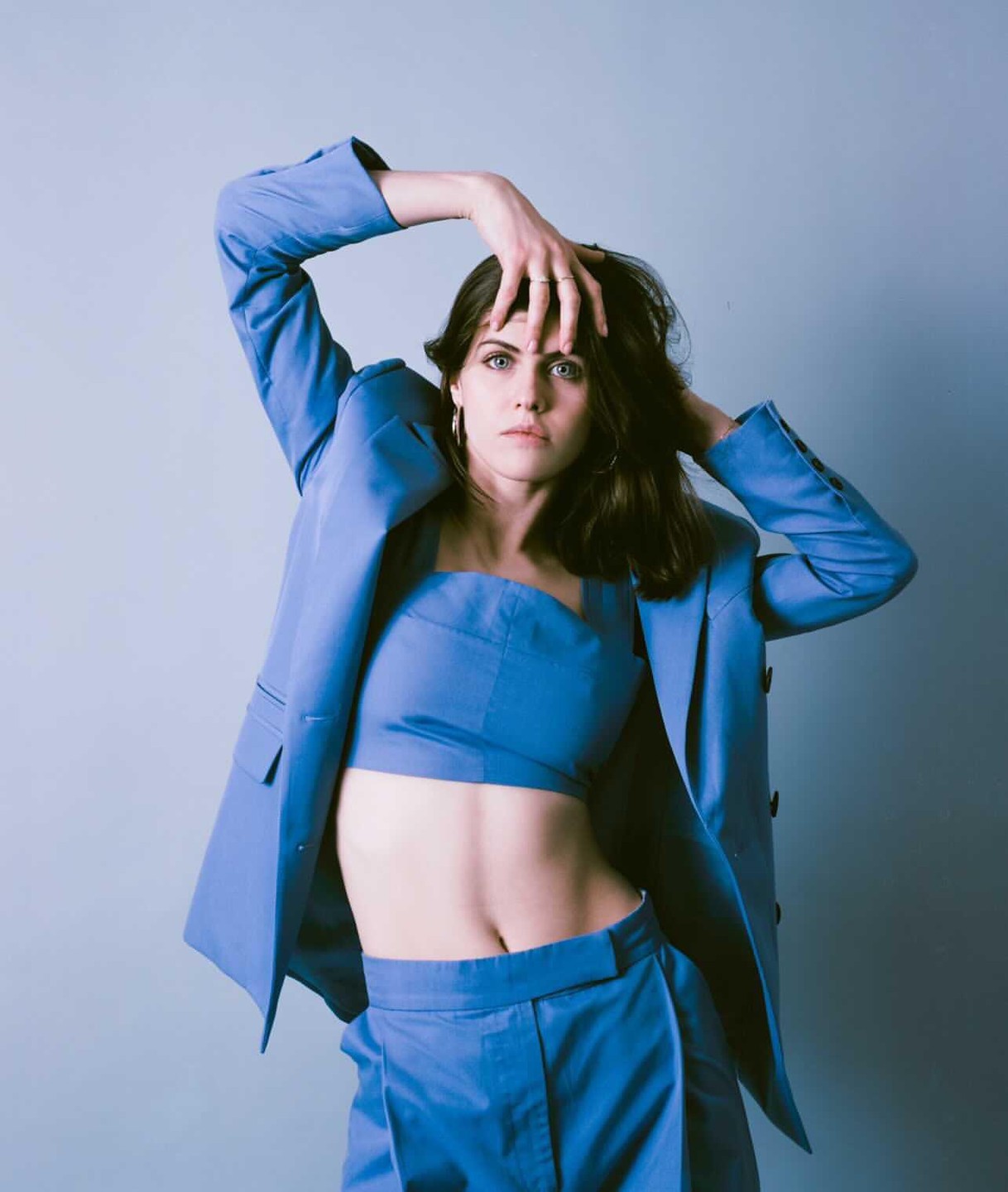 A woman in a blue suit and crop top poses with her hands on her head.