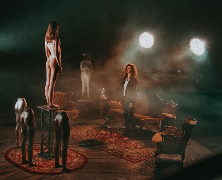 A woman in underwear stands on a pedestal while another woman stands in a room with furniture and mannequins.
