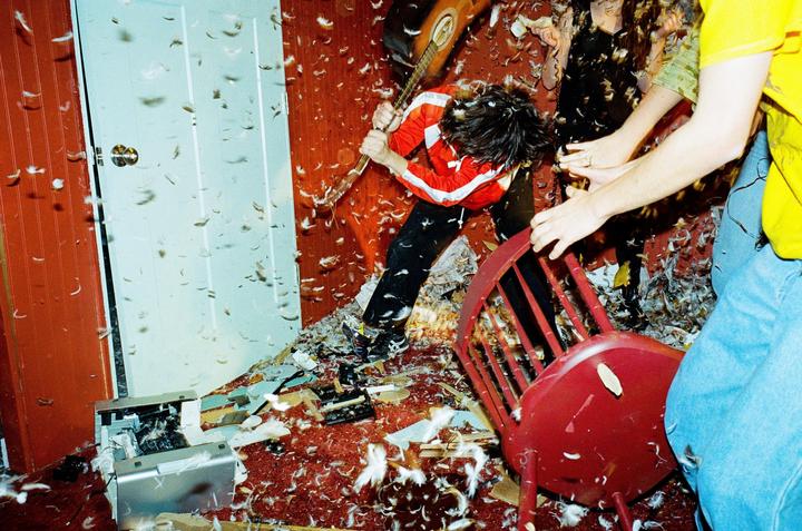 A person in a red jacket smashes a guitar in a room filled with feathers and debris, while another person holds a red chair.