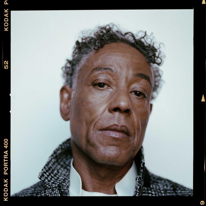 A man with curly hair and a patterned coat looks directly at the viewer.