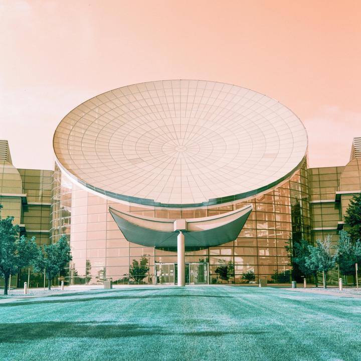 A modern building with a large circular roof and glass facade stands behind a manicured lawn and trees.