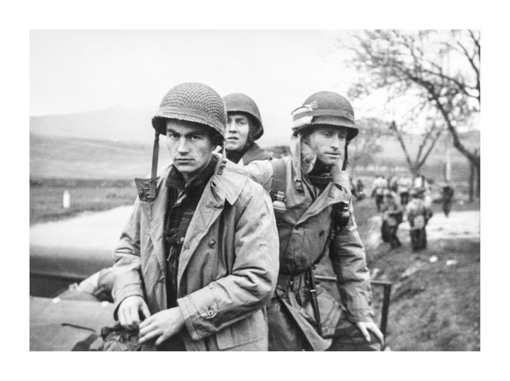 Three soldiers in helmets and coats stand in a line with other soldiers in the background.