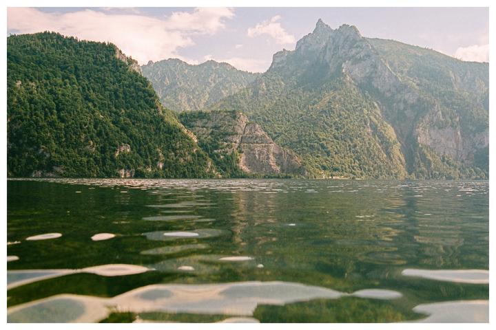 A body of water with ripples on its surface reflects the surrounding mountains covered in trees under a cloudy sky.