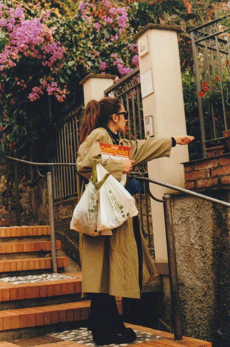 A woman in a trench coat and sunglasses stands on brick steps, holding a magazine and two plastic bags, and pointing towards a gate with flowering bushes in the background.