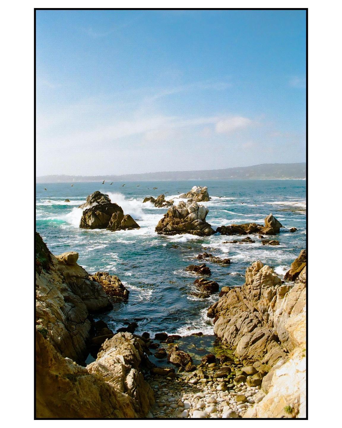 Waves crash against rocky outcroppings along a coastline under a clear blue sky.