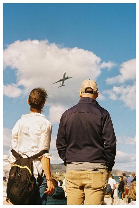 A couple stands with their backs to the viewer, looking up at an airplane flying across a cloudy blue sky.
