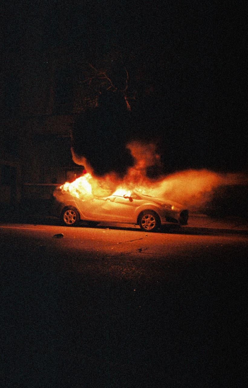 A white car is engulfed in flames on a dark street at night.