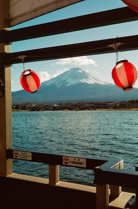 Two red and white lanterns hang in front of a snow-capped mountain and a body of water.