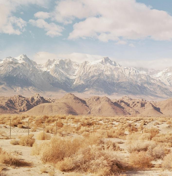 A vast desert landscape features dry brush in the foreground, rocky hills in the middle ground, and snow-capped mountains under a cloudy sky.
