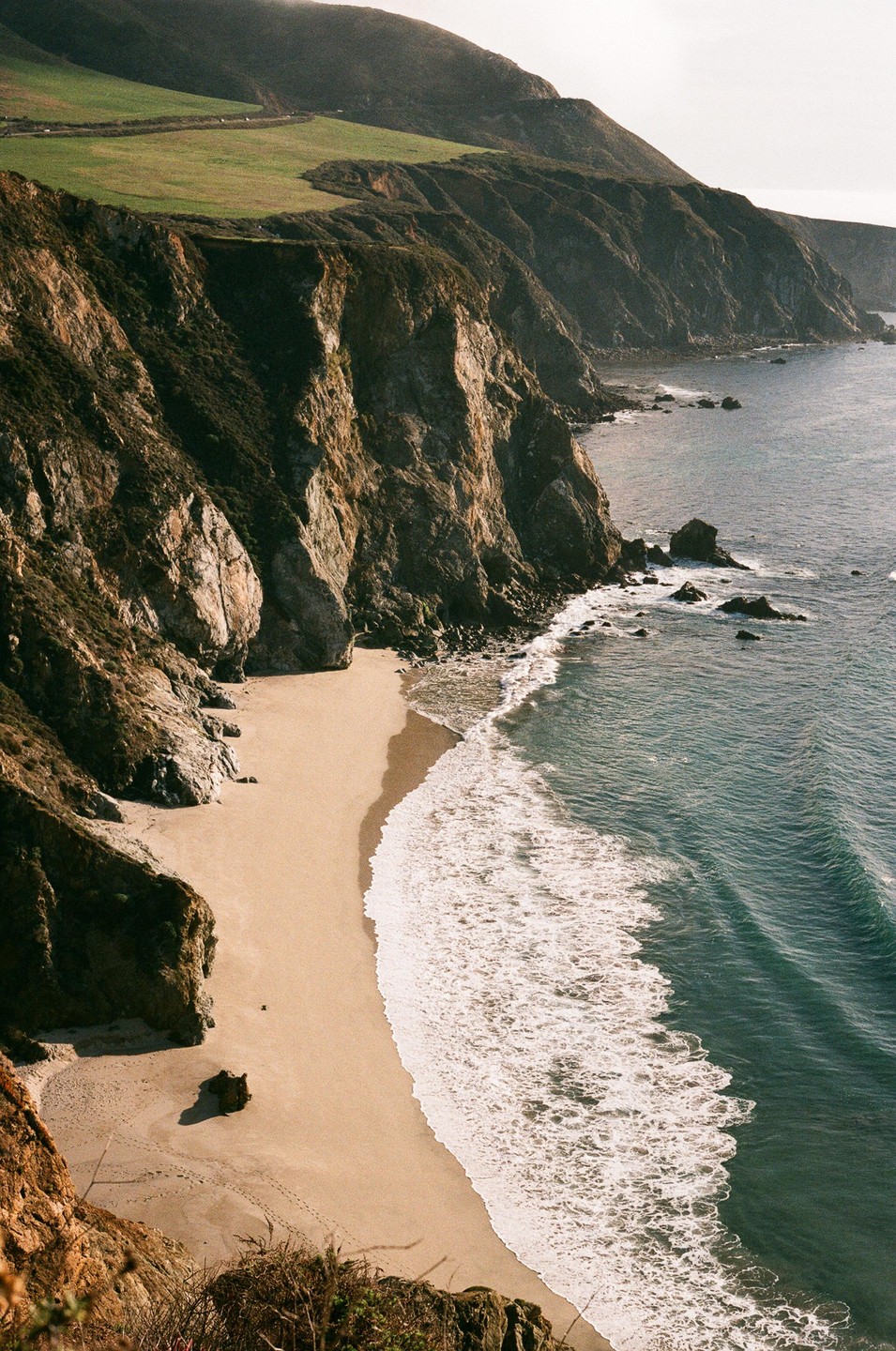 A sandy beach curves along the base of a steep, rocky cliff with green hills above, as waves crash onto the shore.