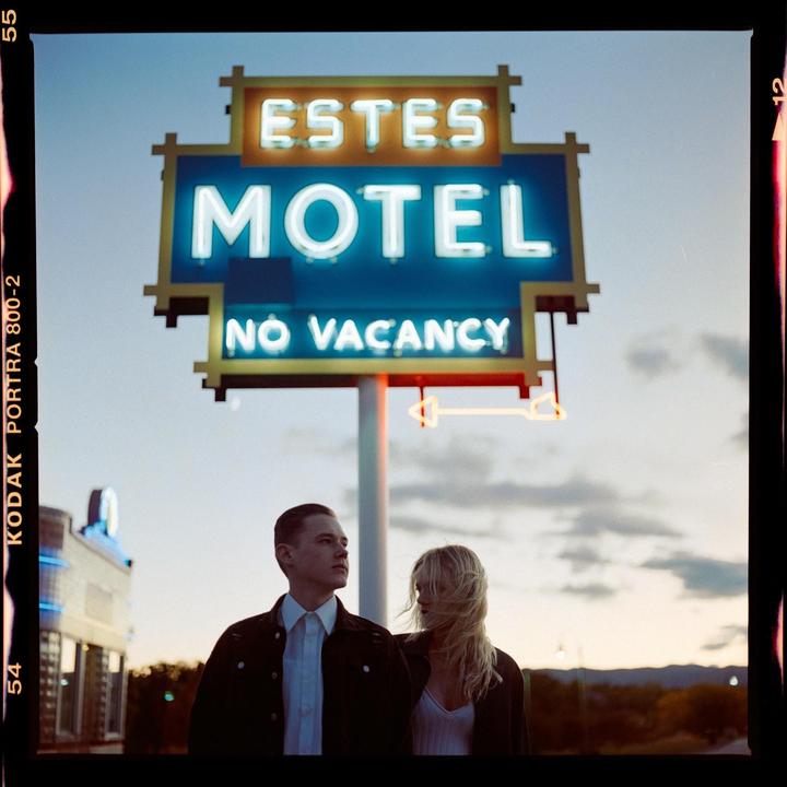 A couple stands in front of a neon motel sign that reads "Estes Motel No Vacancy" at dusk.