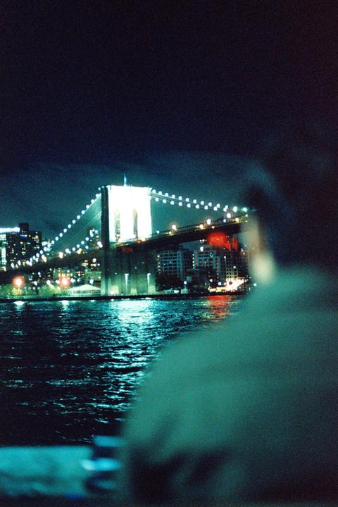 A person looks out over the water at the Brooklyn Bridge lit up at night.