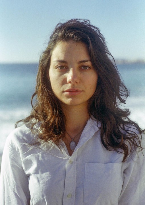 A woman with long, dark hair and a white shirt looks directly at the viewer with the ocean in the background.
