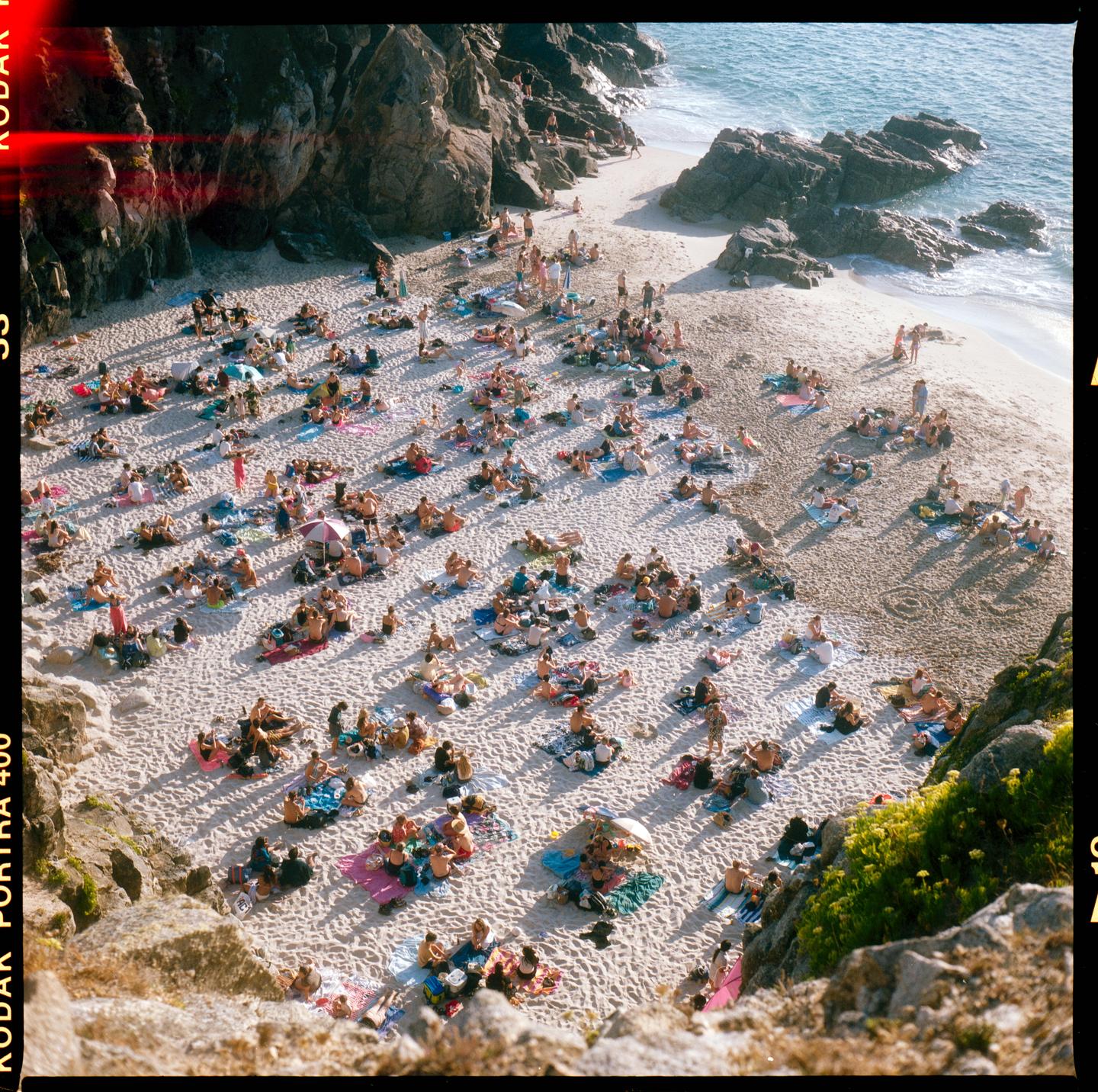 A large crowd of people are sunbathing on a sandy beach next to a rocky coastline.