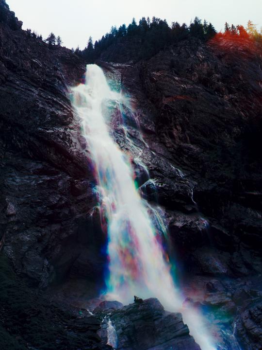 A vibrant rainbow waterfall cascades down a dark, rocky mountain with a forest at the top.