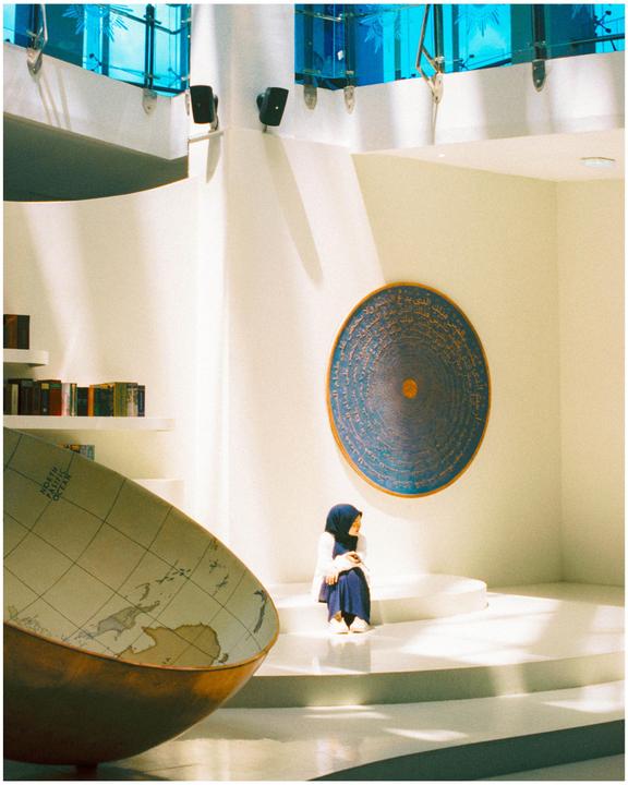 A woman in a hijab sits on steps next to a large globe and a circular artwork on a white wall.