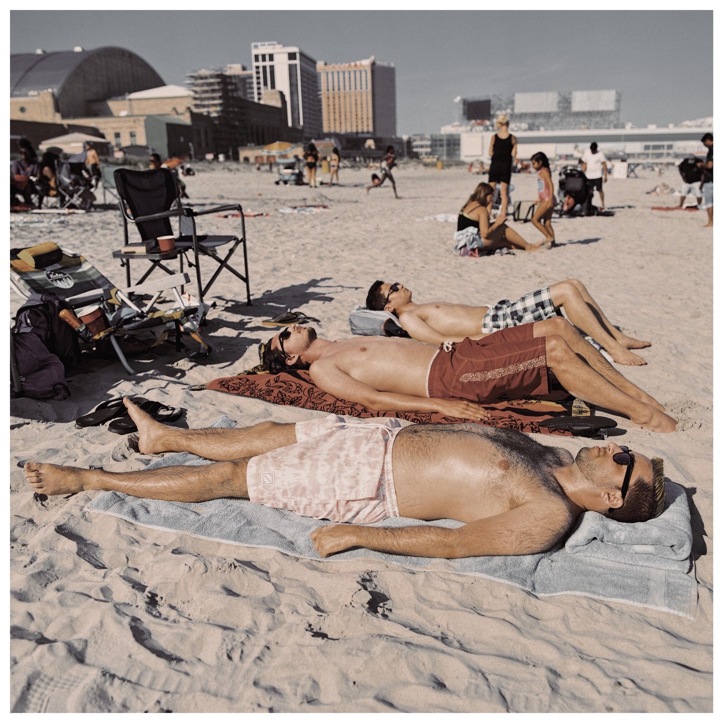 Three men lie on towels on a sandy beach with buildings in the background.