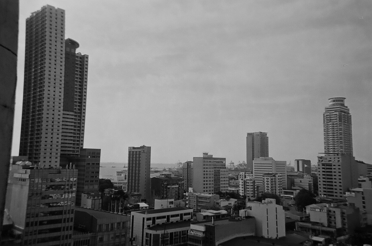 A black and white cityscape shows numerous tall buildings under a cloudy sky, with a body of water and distant ships visible in the background.