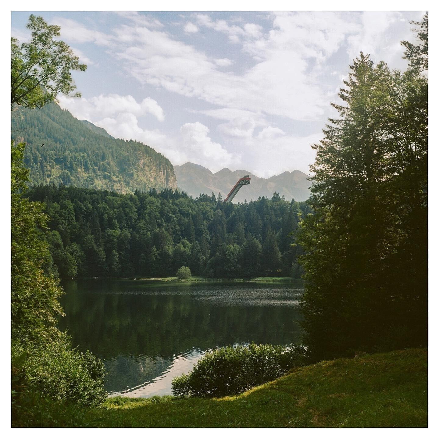A ski jump is visible over a forest and lake, with mountains in the background under a cloudy sky.