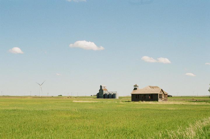 An old wooden building and a grain elevator stand in a green field under a blue sky with scattered clouds.