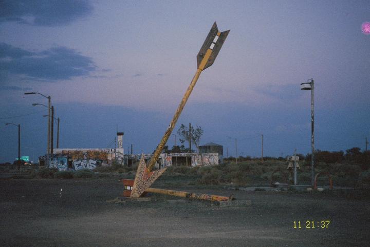 A large, broken arrow sign points towards a graffiti-covered building at dusk.
