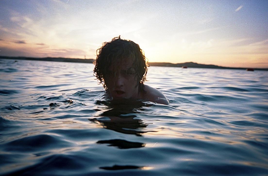 A young man with curly hair is in the water at sunset.
