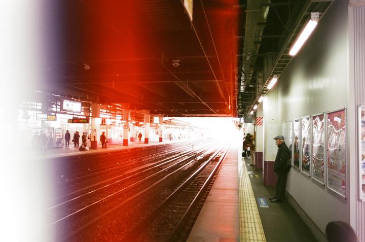 A man stands on a train platform with bright light at the end of the tracks and a red light leak across the top of the image.