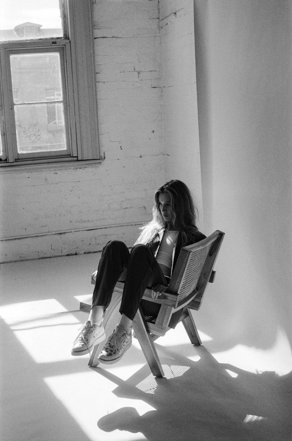 A woman sits in a wooden chair with her legs extended, illuminated by strong window light.