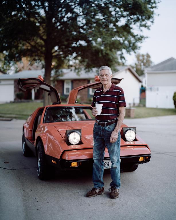 A man stands in front of an orange car with its gull-wing doors open.