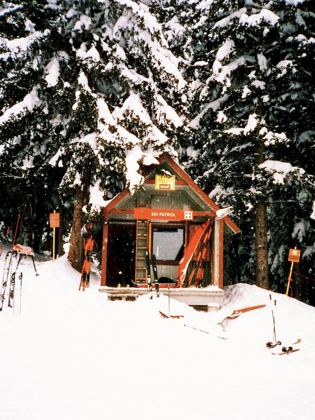 A small ski patrol hut is nestled in a snowy forest, surrounded by skis and ski poles.