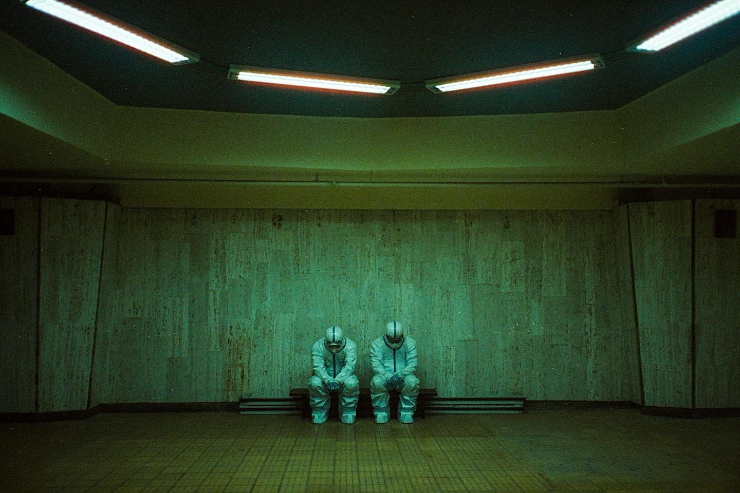 Two people in hazmat suits sit on a bench in a dimly lit, green-hued room.