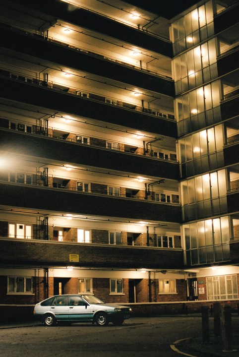 A car is parked in front of a multi-story building with illuminated balconies and windows at night.