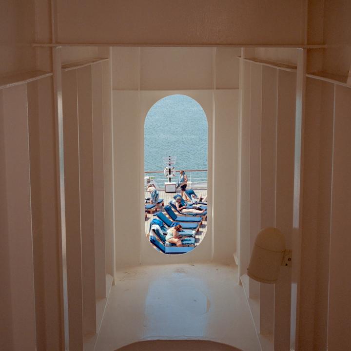 A view through an arched opening shows people relaxing on lounge chairs on a ship deck with the ocean in the background.